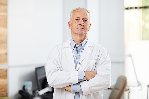 Waist up portrait of experienced senior doctor  while posing in office of modern clinic standing with arms crossed, copy space