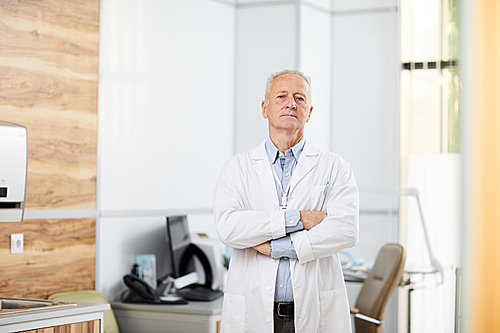 Portrait of experienced senior doctor wearing white coat posing in modern office standing with arms crossed and , copy space