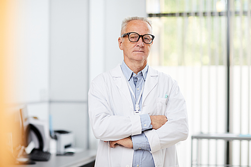Portrait of friendly senior doctor wearing glasses  while posing in office standing with arms crossed, copy space