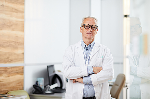 Waist up portrait of senior doctor wearing glasses  while standing with arms crossed in office of modern clinic, copy space