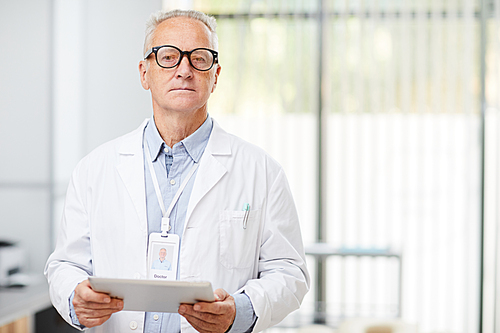Waist up portrait of senior doctor holding digital tablet and  while standing in office of modern clinic
