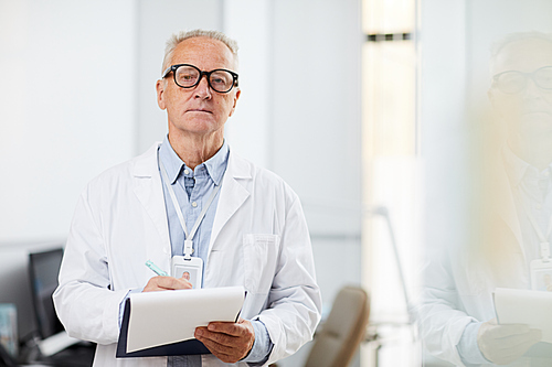 Waist up portrait of senior doctor holding clipboard and looking away pensively while taking notes standing in office, copy space