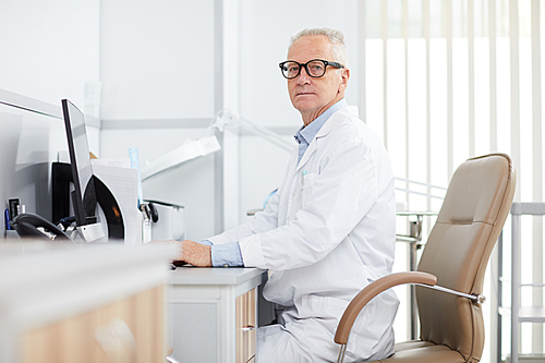 Portrait of senior doctor sitting at desk and  while posing in office of modern clinic, copy space