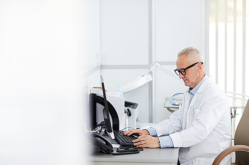 Side view portrait of senior doctor sitting at desk and using computer in office of modern clinic, copy space