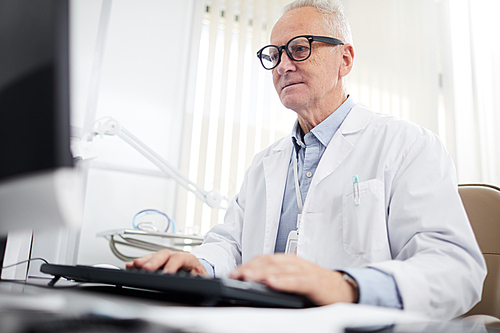 Low angle portrait of senior doctor sitting at desk and using computer in office of modern clinic, copy space