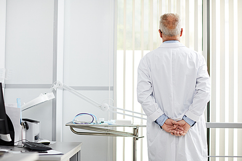Back view portrait of pensive senior doctor looking at window while standing in office of modern clinic, copy space