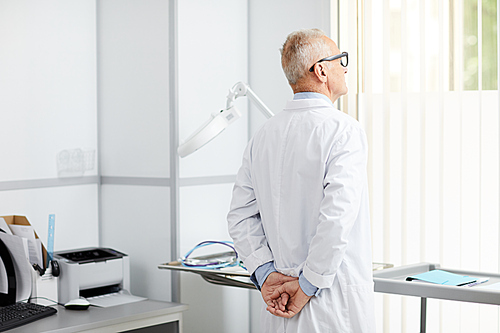 Portrait of pensive senior doctor looking at window while standing in office of modern clinic, copy space
