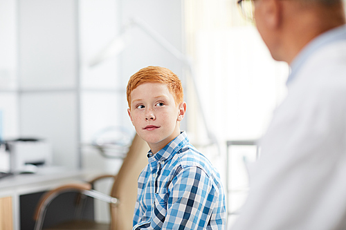 Portrait of cute red haired kid looking at senior doctor during consultation in child healthcare clinic, copy space