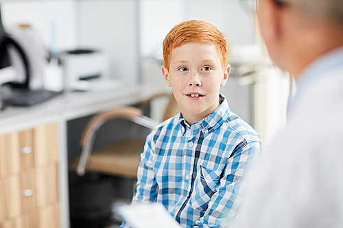 Portrait of cute red haired boy looking at senior doctor during consultation in child healthcare clinic, copy space