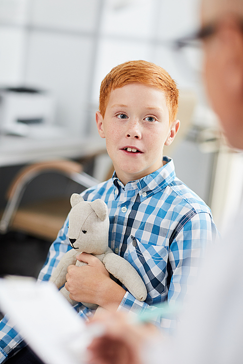 Portrait of cute red haired boy holding bear toy and looking at doctor during consultation in child healthcare clinic