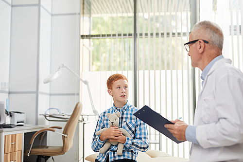 Portrait of cute red haired boy talking to senior doctor during consultation in child healthcare clinic, copy space