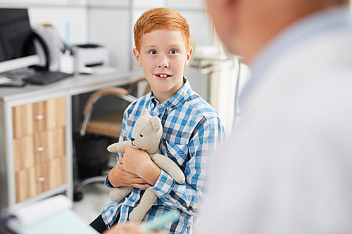 Portrait of cute red haired boy smiling looking at unrecognizable doctor during consultation in child healthcare clinic, copy space