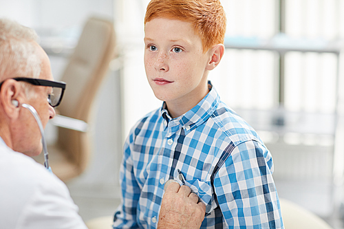 Portrait of senior doctor using stethoscope listening to heartbeat and breathing of cute red haired boy during consultation in child healthcare clinic, copy space