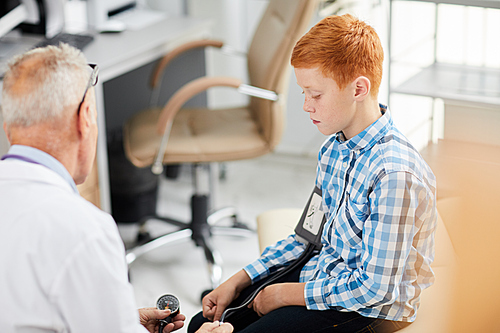 Portrait of senior doctor measuring blood pressure of cute red haired boy during consultation in child healthcare clinic, copy space
