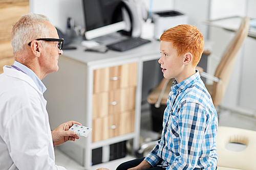 High angle portrait of red haired teenage boy talking to senior doctor sitting in office of modern clinic or hospital, copy space