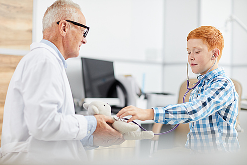 Portrait of cute red haired boy listening to heartbeat of plush bear using stethoscope during consultation with senior doctor in child clinic, copy space