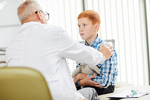 Portrait of cute red haired boy in doctors office looking at senior physician during consultation in child clinic, copy space