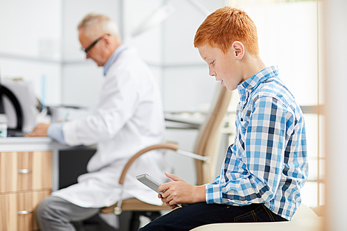 Side view portrait of red haired teenage boy using digital tablet in waiting room at doctors office, copy space