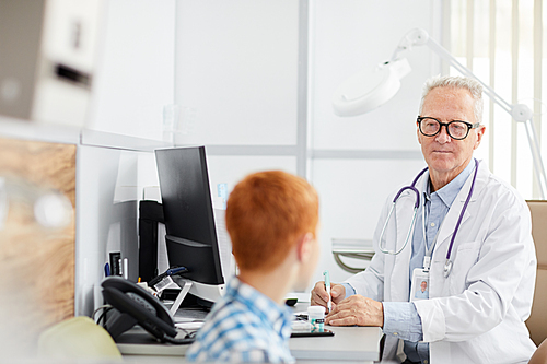 Portrait of white-haired senior doctor talking to little patient during consultation in child healthcare clinic, copy space