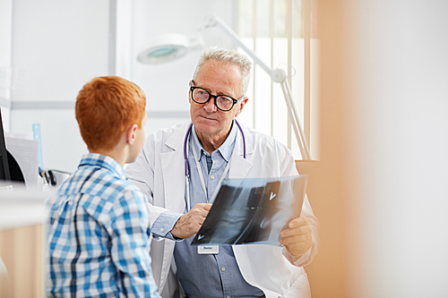 Portrait of white-haired senior doctor holding x ray image while talking to little patient during consultation in child healthcare clinic, copy space