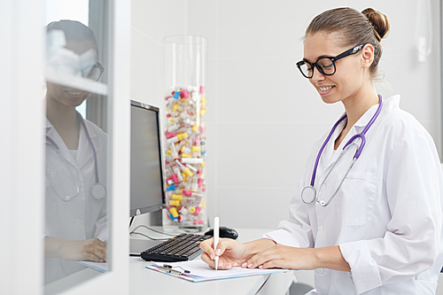 Side view portrait of smiling female doctor writing on clipboard while sitting at desk in office, copy space
