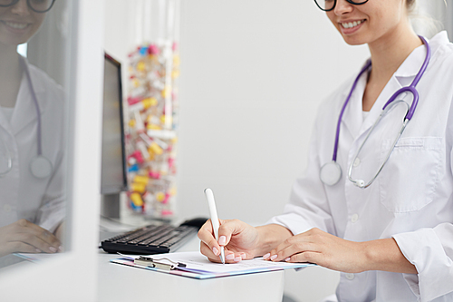Mid-section portrait of smiling female doctor writing on clipboard while sitting at desk in office, copy space