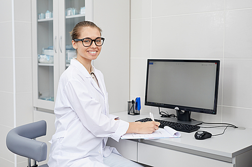 Portrait of pretty female doctor wearing glasses smiling cheerfully at camera while taking notes at desk in office, copy space