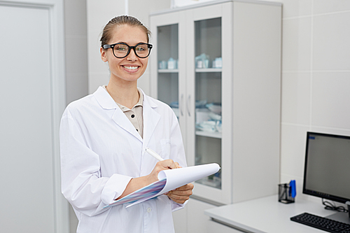 Waist up portrait of pretty female doctor holding clipboard and smiling cheerfully at camera while posing in office, copy space