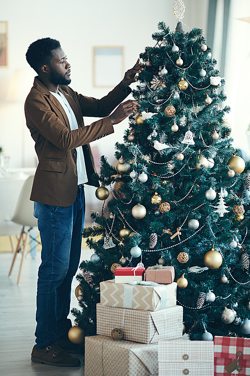 Full length portrait of modern African-American man decorating Christmas tree at home, copy space