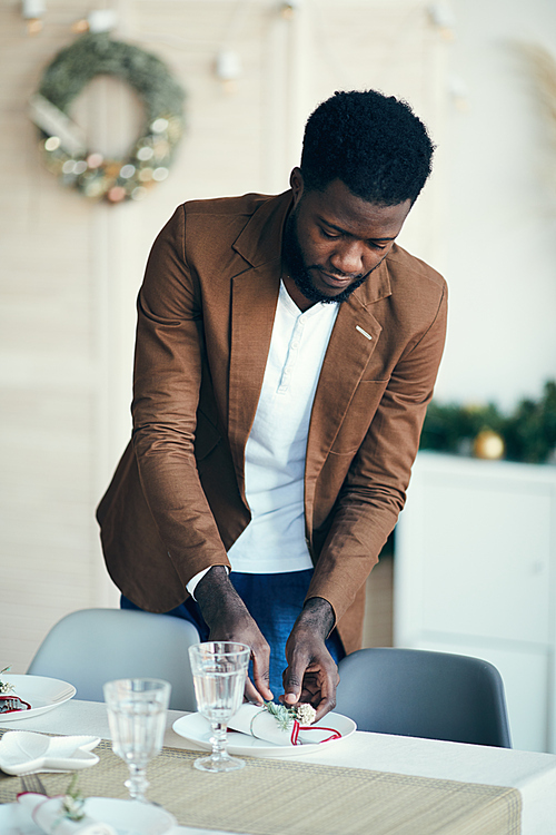 Portrait of modern African-American man preparing table setting while decorating dining room for Christmas party at home