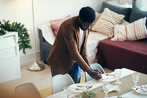 High angle portrait of young African-American man preparing table setting while decorating dining room for Christmas party, copy space