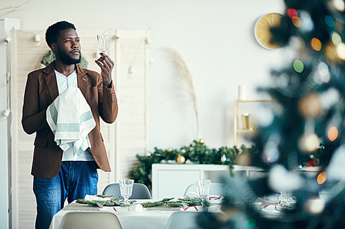 Portrait of young African-American man wiping crystal glasses while setting dinner table for Christmas party, copy space
