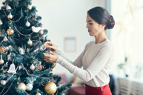 Side view portrait of elegant young woman decorating Christmas tree at home, copy space
