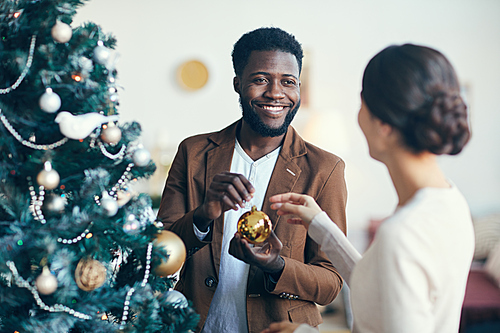 Waist up portrait of modern mixed-race couple decorating Christmas tree together at home, copy space