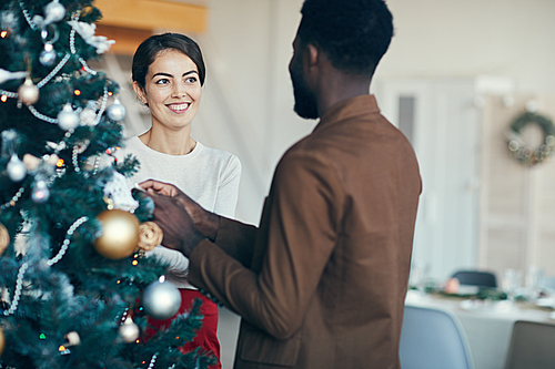 Portrait of modern mixed-race couple decorating Christmas tree together at home, copy space