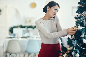 Portrait of elegant young woman speaking by phone while decorating Christmas tree at home, copy space