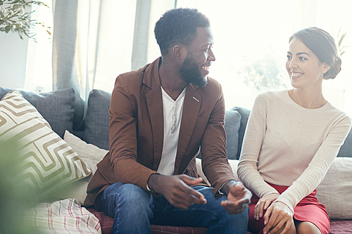 Portrait of mixed-race couple smiling at each other while talking sitting on cozy sofa at home, copy space