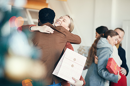 Group of modern young people exchanging gifts during Christmas party, focus on smiling woman hugging friend in foreground, copy space