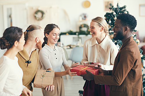 Waist up portrait of cheerful young people exchanging presents during Christmas party in cozy interior, copy space