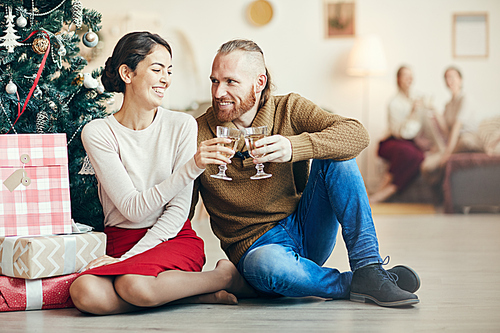 Full length portrait of modern adult couple clinking champagne glasses sitting by Christmas tree at home, copy space