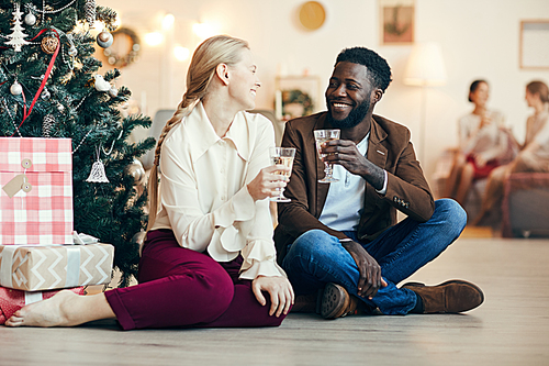 Full length portrait of mixed-race couple clinking champagne glasses sitting by Christmas tree at home, copy space