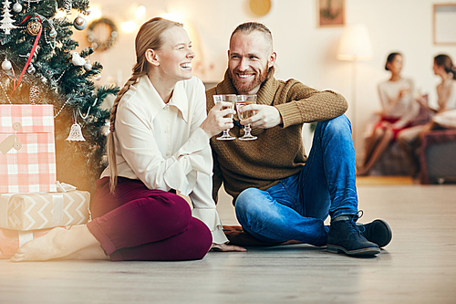 Full length portrait of happy couple clinking champagne glasses sitting by Christmas tree at home, copy space