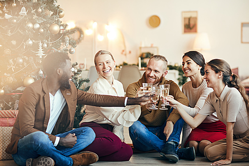 Full length portrait of cheerful friends clinking champagne glasses sitting by Christmas tree during party, copy space