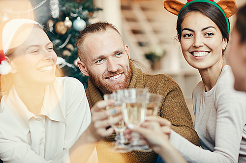 Portrait of friends clinking champagne glasses sitting by Christmas tree during party, copy space