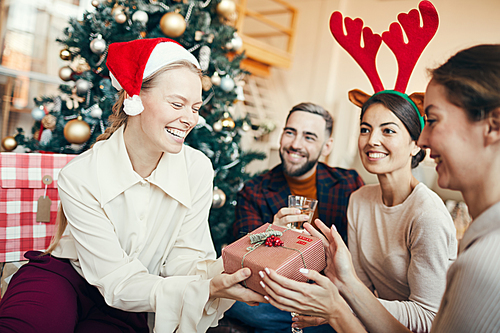 Group of cheerful adult people exchanging gifts during Christmas party, focus on happy young woman in foreground