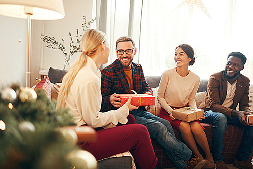 Group of elegant adult people exchanging presents on Christmas, focus on bearded man smiling happily holding red gift box, copy space