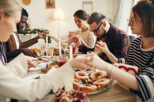 Multi-ethnic group of people sitting at dining table on Christmas and joining hands in prayer, copy space