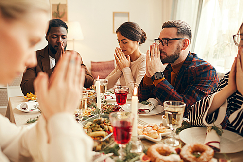 Multi-ethnic group of elegant adult people praying sitting at dinner table during Christmas banquet