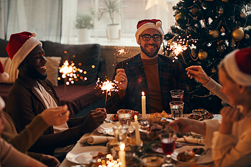 Group of cheerful people holding sparklers while enjoying Christmas dinner at home