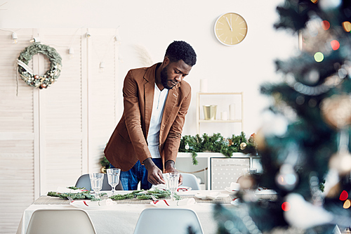 Portrait of young African-American man setting table for Christmas banquet and decorating beautiful dining room, copy space